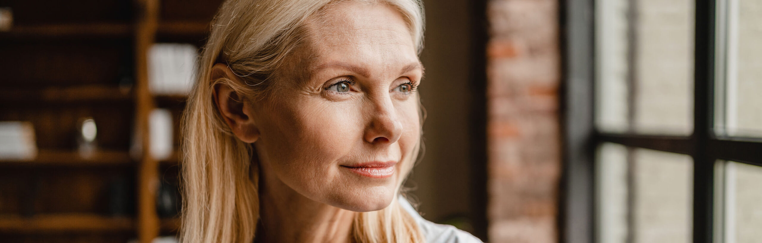 A middle-aged woman with blonde hair looks thoughtfully out of a window, soft natural light illuminating her face as she reflects on her experience with domestic violence.